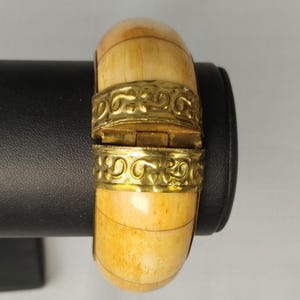 May include: A cream-coloured, hinged bangle bracelet with ornate gold-coloured metal bands. The bracelet has a barrel shape and a clasp that opens to reveal the interior. The metal bands feature a repeating scroll design.