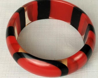 May include: A bold, round bangle bracelet with a striking pattern of alternating red and black stripes. The bracelet is made of a glossy material and has a smooth, polished finish. The design is eye-catching and suitable for everyday wear.