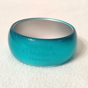 May include: A teal-colored, rounded bangle bracelet with the engraved text "OMNIA PARAÍBA". The bracelet has a glossy finish and a white interior. The bangle is a fashion accessory.