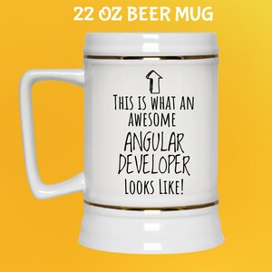 May include: A white 22 oz beer mug with gold trim and a handle. The mug has the text "THIS IS WHAT AN AWESOME ANGULAR DEVELOPER LOOKS LIKE!" in black. The mug is on a yellow background.