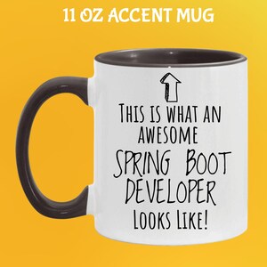 May include: White 325 ml accent mug with a black handle and rim. The mug features the text "THIS IS WHAT AN AWESOME SPRING BOOT DEVELOPER LOOKS LIKE!" in black, with an arrow graphic. The mug is on a yellow background.