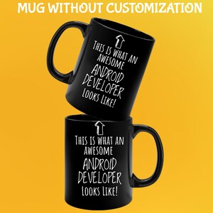 K&ouml;nnte beinhalten: Zwei schwarze Keramikbecher, die &uuml;bereinander gestapelt sind. Jeder Becher hat einen gro&szlig;en Henkel und wei&szlig;en Text, der lautet: "THIS IS WHAT AN AWESOME ANDROID DEVELOPER LOOKS LIKE!" mit einer Pfeilgrafik nach oben. Der Hintergrund ist gelb.