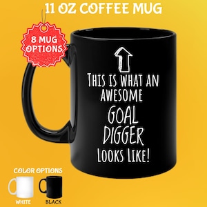 May include: Black 11 oz coffee mug with the text "THIS IS WHAT AN AWESOME GOAL DIGGER LOOKS LIKE!" in white. The mug has a black handle and a white arrow graphic. Color options include white and black mugs.