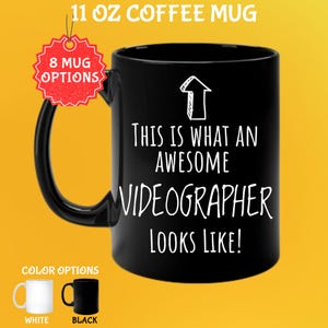 May include: Black 11 oz coffee mug with the text "THIS IS WHAT AN AWESOME VIDEOGRAPHER LOOKS LIKE!" in white. The mug has a black handle and a white arrow graphic. Color options include white and black mugs.
