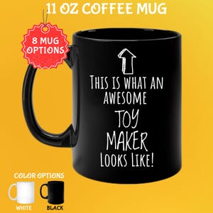 May include: Black 11 oz coffee mug with the text "THIS IS WHAT AN AWESOME TOY MAKER LOOKS LIKE!" in white. The mug has a black handle and a white arrow graphic. The image also shows white and black color options.