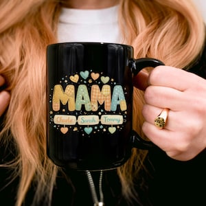 Könnte beinhalten: Eine schwarze Keramik-Tasse mit dem Wort "MAMA" in einem farbenfrohen, gemusterten Design. Die Tasse zeigt auch die Namen "Charlie", "Sarah" und "Tommy" unter dem Wort "MAMA", mit Herz- und Sternakzenten.