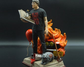 May include: A figurine of a man holding an open book with the text "We All Float Down Here" on his shirt. He is standing on a book with a red balloon, a paint can, and a cat figurine. The scene is set against a backdrop of flames.