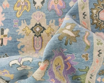 May include: Close-up of a blue rug with a detailed floral pattern in shades of pink, purple, yellow, and beige. The rug has a folded section, revealing the texture and design of the fabric. The rug's design is intricate and detailed.
