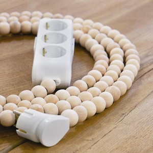 Op de afbeelding: Een witte stekkerdoos met drie stopcontacten en een bijpassende stekker, verbonden door een snoer van lichtgekleurde houten kralen. De kralen zijn in een decoratief, gebogen patroon gerangschikt. De stekkerdoos is ontworpen voor thuis of op kantoor.