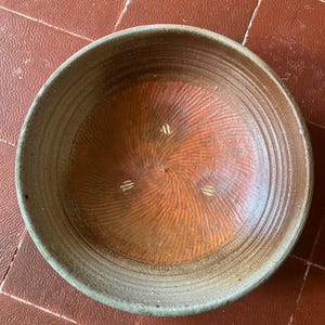 Op de afbeelding: Een handgemaakte keramische kom met een breed, ondiep ontwerp. De binnenkant heeft een roodbruine kleur met een spiraalpatroon, terwijl de rand en de buitenkant een gedempt groenbruin zijn. De kom is geschikt voor serveren of decoratie.