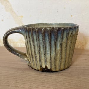Op de afbeelding: Handgemaakte keramische mok met een geribbelde buitenkant. De mok heeft een handvat en een uniek glazuur met tinten blauw, groen en bruin. De mok is ontworpen voor warme dranken.
