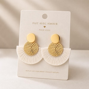 Peut inclure: Boucles d'oreilles dorées avec un dessus circulaire et un bas en forme de ventilateur texturé en franges blanches. Les boucles d'oreilles sont présentées sur une carte blanche avec le texte français : "FAIT AVEC AMOUR POUR VOUS" et "BIJOUX QUI SUBLIMENT CHAQUE INSTANT."