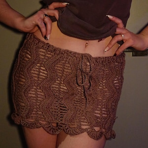 May include: A brown, crocheted mini skirt with a drawstring waist and scalloped hem. The skirt has an open weave design. The model is wearing a brown ribbed top.