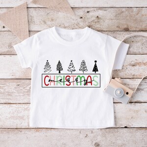 Könnte beinhalten: Weißes T-Shirt mit einem Motiv aus fünf stilisierten Weihnachtsbäumen und dem Text "merry christmas" in roten, grünen und rosa Buchstaben.