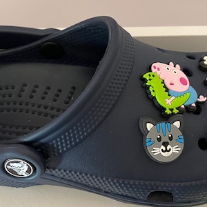 May include: A navy blue clog shoe decorated with cartoon character charms. The charms feature a pink pig with a green dinosaur, a grey cat, a giraffe, and a dragonfly. The shoe has a textured surface and a logo on the side.