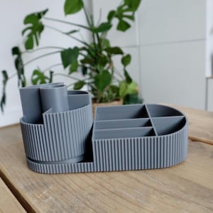 May include: A grey desk tidy with several compartments and a cylindrical section for pens or pencils. The organiser has a ribbed texture and sits on a wooden surface. A green plant is visible in the background.
