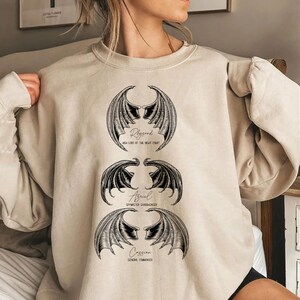 May include: A beige sweatshirt with a graphic print of three sets of wings with the names Rhysand, Azriel, and Cassian and their titles from the book A Court of Thorns and Roses.
