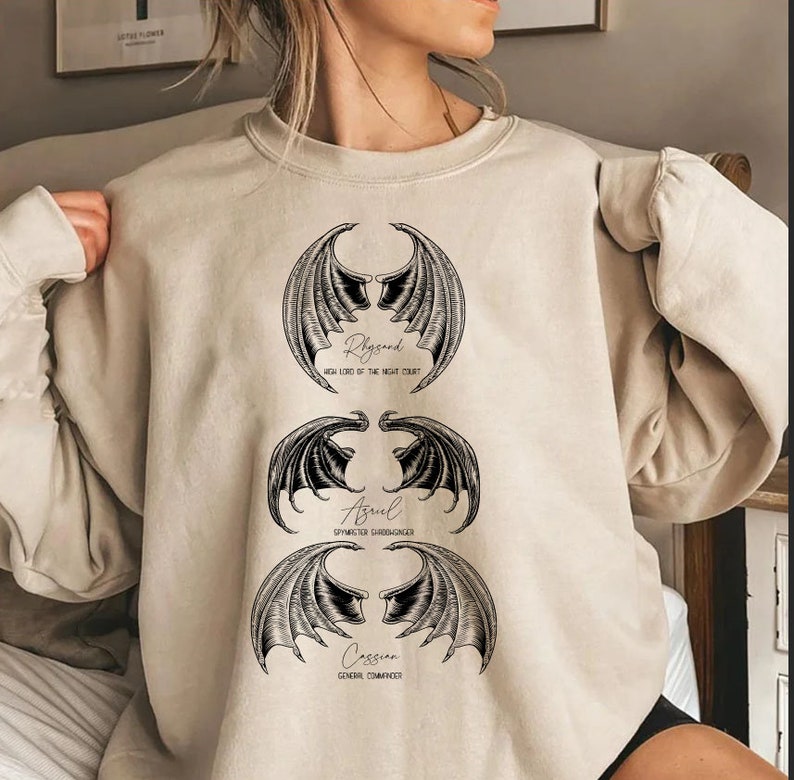May include: A beige sweatshirt with a graphic print of three sets of wings with the names Rhysand, Azriel, and Cassian and their titles from the book A Court of Thorns and Roses.