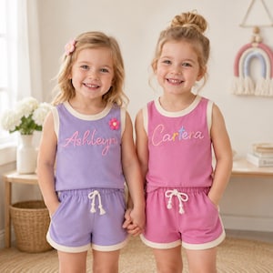 May include: Two young girls wearing matching lavender and pink tank tops and shorts sets. The lavender set has the name "Ashleyn" embroidered on the front with a flower, while the pink set says "Cartera". Both sets have white trim.