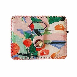 May include: A pink leather card holder with a floral print and a silver snap closure. The print features abstract flowers in shades of red, orange, green, and blue.