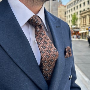 May include: A navy blue double-breasted suit with a white dress shirt and a patterned brown tie. A matching pocket square is visible in the chest pocket. The suit has silver buttons.