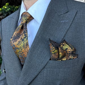 Op de afbeelding: Een grijs dubbelbreasted pak met een lichtblauw overhemd en een stropdas en pochet met patroon. De stropdas en pochet hebben een zwart en oranje palmbladontwerp. Het pak heeft zilveren knopen.
