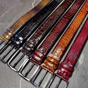 Op de afbeelding: Een collectie van zeven leren riemen in verschillende kleuren, waaronder bruin, zwart, bordeauxrood en rood. Elke riem heeft een getextureerd, reliëfpatroon en een zilverkleurige rechthoekige gesp. De riemen zijn diagonaal gerangschikt en tonen hun verschillende tinten.