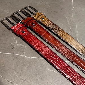 May include: Four leather belts in shades of red, brown, and tan. Each belt has a textured, crocodile-like pattern and a silver-colored buckle. The belts are arranged diagonally, showcasing their varying colors and textures.