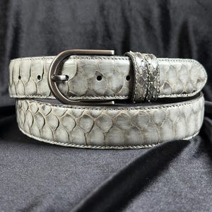 Op de afbeelding: Een lichtgrijze riem met een gestructureerd slangenleerpatroon met grote, overlappende schubben. De riem is voorzien van een donker metalen gesp en een bijpassende riemlus van slangenleer, met een consistent schubbenpatroon over de hele band.