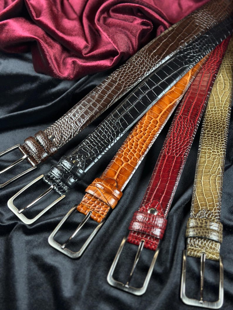 May include: An assortment of leather belts in various colors, including brown, black, orange, red, and olive green. Each belt features a textured, crocodile-like pattern and a silver-colored buckle. The belts are displayed on a black fabric background.
