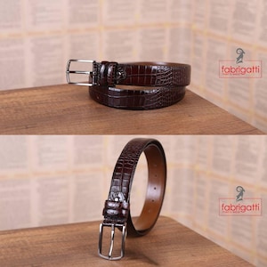 May include: Two brown leather belts with a crocodile skin texture are displayed on a wooden surface. The belts have silver-colored buckles and are from the brand Fabrigatti. The belts are a rich, dark brown color.
