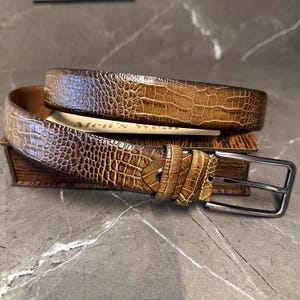 May include: A brown leather belt with a crocodile skin texture. The belt has a silver-colored buckle and is resting on a small wooden box. The words "Men's wear" are visible on a piece of paper.