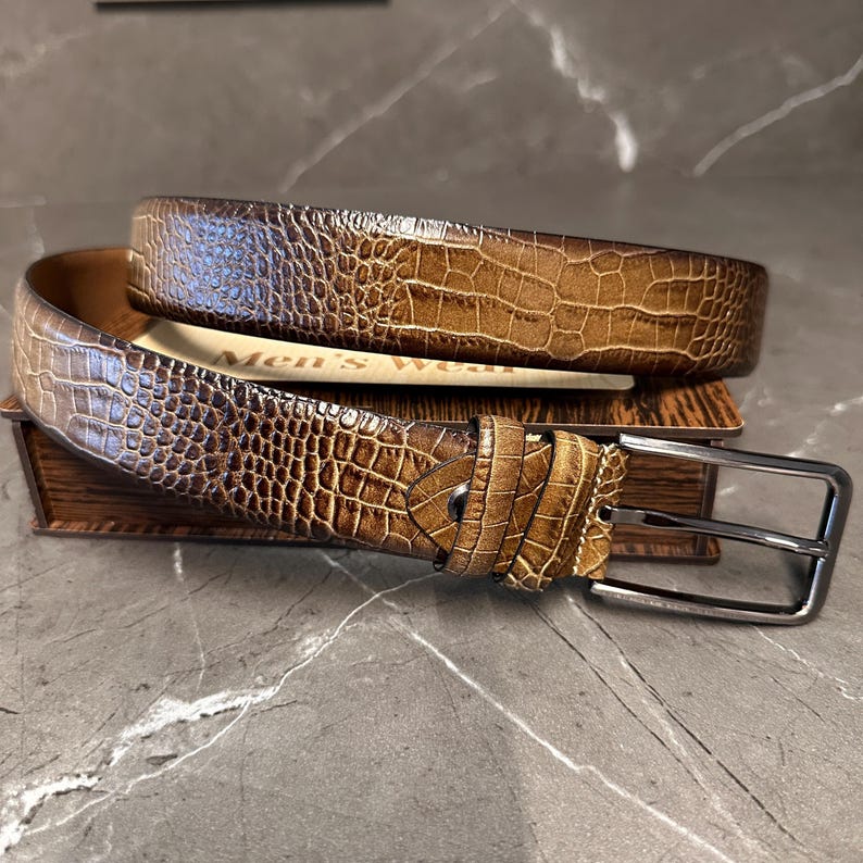May include: A brown leather belt with a crocodile skin texture. The belt has a silver-colored buckle and is resting on a small wooden box. The words "Men's wear" are visible on a piece of paper.