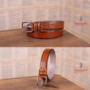 May include: Two brown leather belts with a crocodile texture are displayed on a wooden surface. The belts have silver-colored buckles and are arranged in two different orientations. The brand name "fabrigatti men's wear" is visible in the image.
