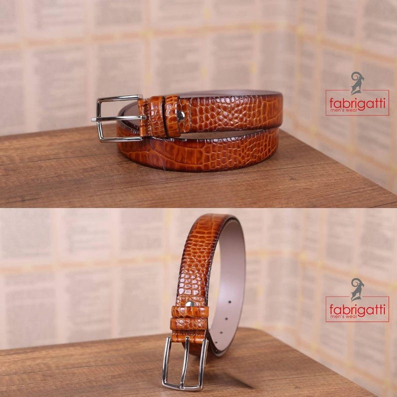 May include: Two brown leather belts with a crocodile texture are displayed on a wooden surface. The belts have silver-colored buckles and are arranged in two different orientations. The brand name "fabrigatti men's wear" is visible in the image.