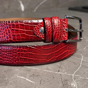 May include: A red belt with a textured, crocodile-like pattern. The belt has a silver-colored buckle and a matching belt loop. The leather belt is a rich, deep red color.
