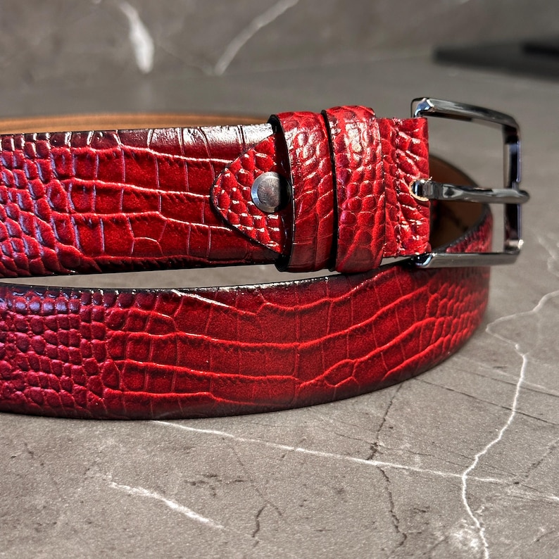 May include: A red belt with a textured, crocodile-like pattern. The belt has a silver-colored buckle and a matching belt loop. The leather belt is a rich, deep red color.