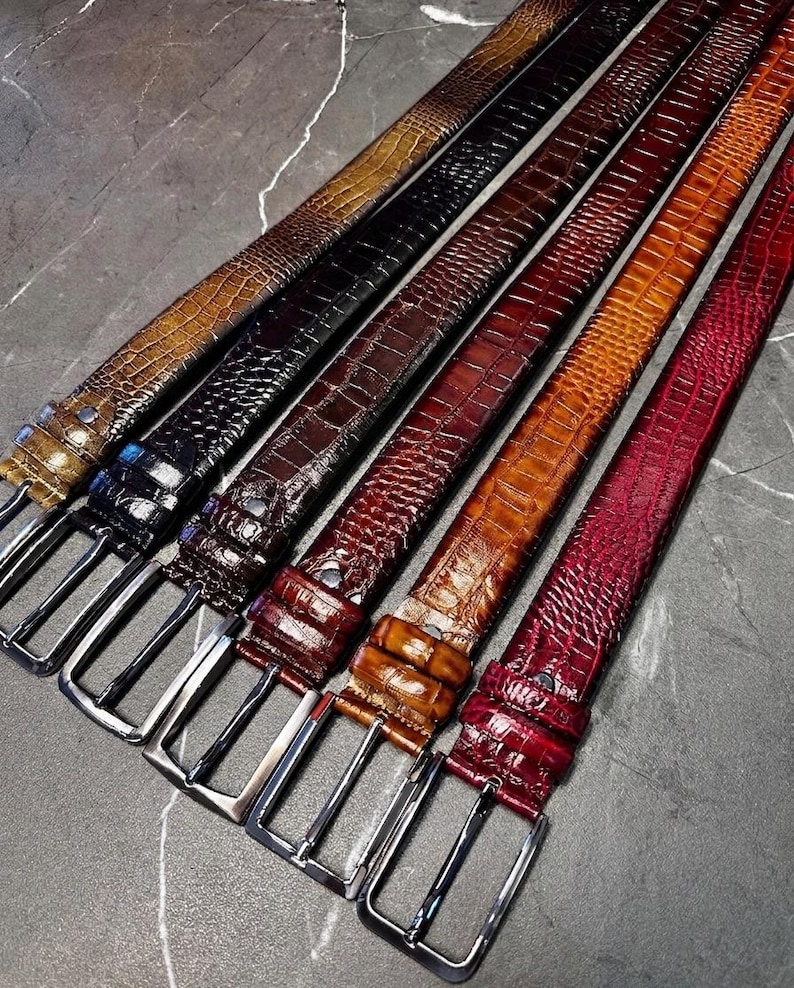 May include: A collection of seven leather belts in various colors, including brown, black, burgundy, and red. Each belt features a textured, embossed pattern and a silver-colored rectangular buckle. The belts are arranged diagonally, showcasing their different hues.