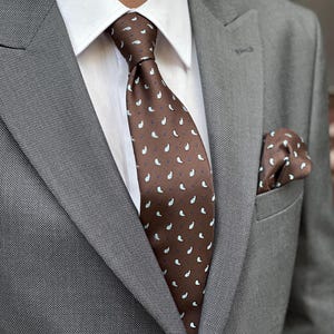 Puede incluir: Una corbata marrón estampada y un pañuelo de bolsillo a juego se combinan con un traje gris y una camisa blanca. La corbata presenta un patrón repetido de formas de lágrima blancas y azul claro.
