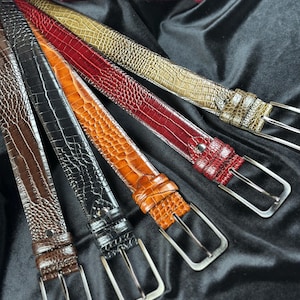May include: A collection of leather belts in various colors, including brown, black, orange, red, and olive green. Each belt features a textured, embossed pattern and a silver-toned buckle. The belts are arranged on a black fabric background.