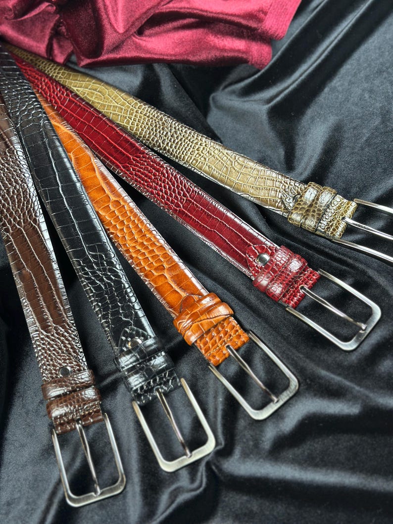 May include: A collection of leather belts in various colors, including brown, black, orange, red, and olive green. Each belt features a textured, embossed pattern and a silver-toned buckle. The belts are arranged on a black fabric background.
