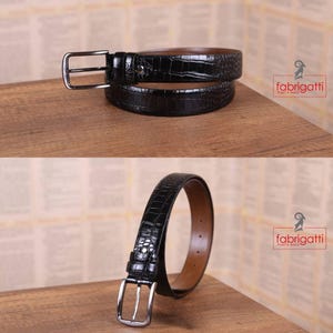 May include: Black leather belt with a crocodile skin texture and a silver-colored buckle. The belt is displayed in two arrangements, showcasing its design and details. The brand name "fabrigatti men's wear" is visible in the image.