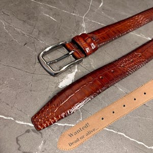 May include: A brown leather belt with a crocodile texture and a silver buckle. The belt has a light brown interior with the text "Wanted! Dead or alive..." embossed on the end. The belt is displayed on a gray marble surface.
