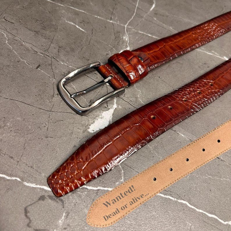 May include: A brown leather belt with a crocodile texture and a silver buckle. The belt has a light brown interior with the text "Wanted! Dead or alive..." embossed on the end. The belt is displayed on a gray marble surface.
