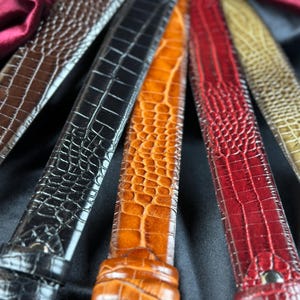 May include: Close-up of several leather belts in various colors, including brown, black, orange, and red. The belts have a textured, crocodile-like pattern and are arranged in a fan shape, showcasing their different hues and designs.