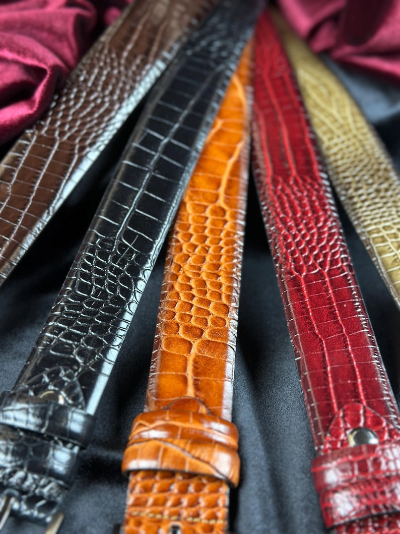 May include: Close-up of several leather belts in various colors, including brown, black, orange, and red. The belts have a textured, crocodile-like pattern and are arranged in a fan shape, showcasing their different hues and designs.