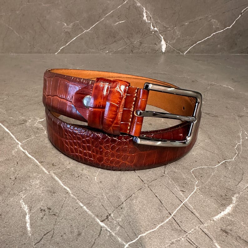 May include: A brown leather belt with a textured, crocodile-like pattern. The belt has a silver-colored buckle and a matching belt loop. The leather is a rich, reddish-brown color, and the belt is rolled up.