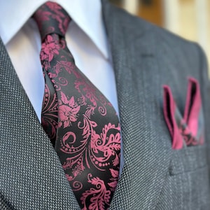May include: A grey suit with a white shirt and a black tie featuring a pink floral design. A matching pink pocket square is visible. The suit has a textured pattern. The image focuses on the upper torso.