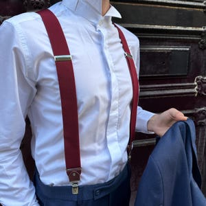 May include: A pair of burgundy suspenders with silver clips and a leather strap. The suspenders are worn over a white button-down shirt and blue pants.
