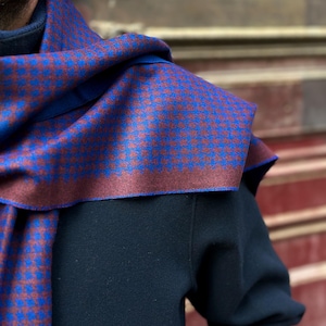 May include: A blue and burgundy patterned scarf draped over a dark blue sweater. The scarf features a repeating pattern of small blue squares on a burgundy background. The scarf's border is a solid burgundy color.